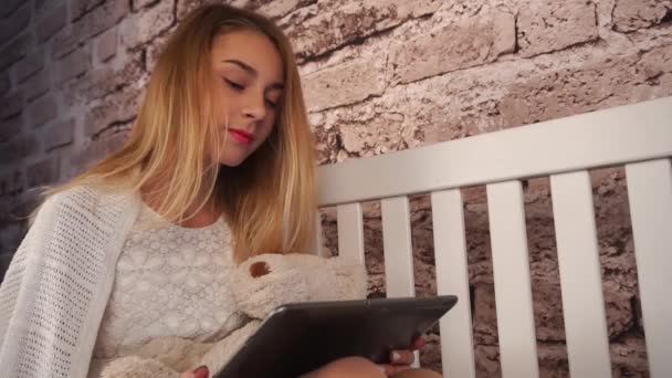 Beautiful girl is sitting on the white bench. She hugging a teddy bear and chatting with friends using the tablet. Pretty face. — Stock Video