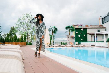 Çekici, sevimli, güzel, yumuşak, güzel Afrikalı kadın elbise çizgili ve siyah şapka kafasına. Avrupa'nın lüks otelin havuzu yakınındaki yürüyüş modeli. İyi bir hava, Rüzgar