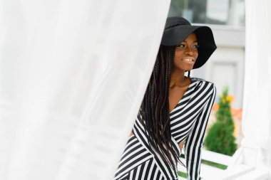 Çekici kadın siyah şapka ve çizgili elbise. Beyaz etrafında ışık sunblind. Model çit oturan ve gülümseyen.