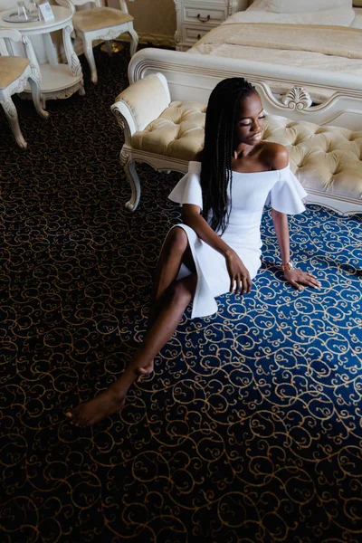 Attraente donna di colore sul pavimento in hotel di lusso. belle gambe e corpo stupendo, occhi chiusi. Abito bianco con spalle nude. Mobili classici, poltrone, divano, letto . — Foto Stock