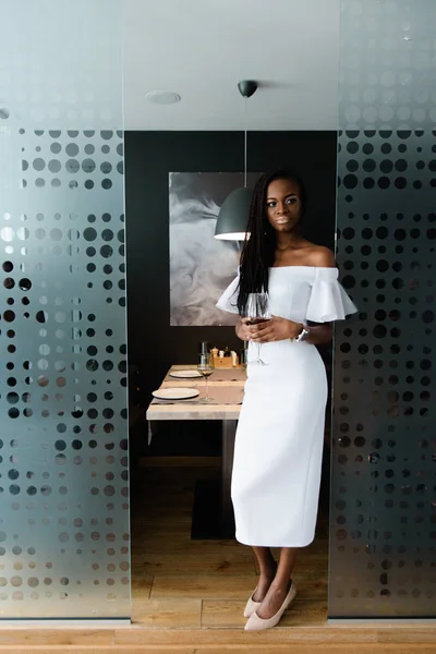 Jeune belle femme africaine séjournant dans un restaurant au design moderne. Elle porte une robe blanche midi avec les épaules nues et a un verre de vin rouge à la main . — Photo