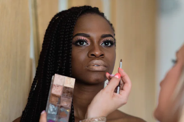 Maskenbildnerin, die afrikanisches Model zum Shooting vorführt. Sie verwendet eine Palette von Grundlagen für Make-up. schwarzes Modell mit langen dunklen Haaren. — Stockfoto