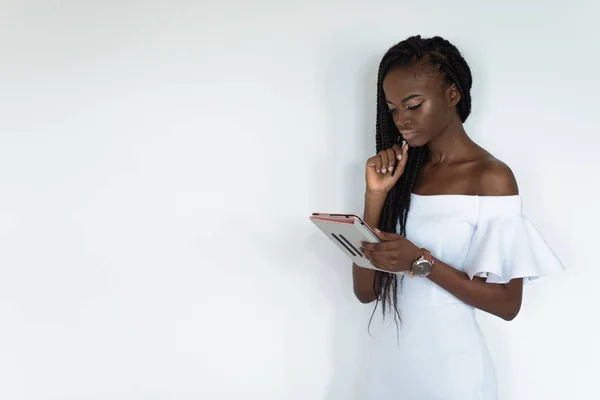 Sexy model met zwarte huid is voor een witte muur staan en chatten op internet met behulp van haar tablet. Meisje is het dragen van witte jurk met blote schouders en horloge op haar hand — Stockfoto