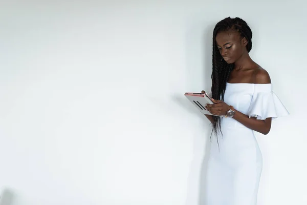 Sexy jolie modèle photo avec peau noire est debout devant un mur blanc et bavarder sur Internet à l'aide de sa tablette. Fille porte longue robe blanche avec les épaules nues et regarder sur sa main — Photo