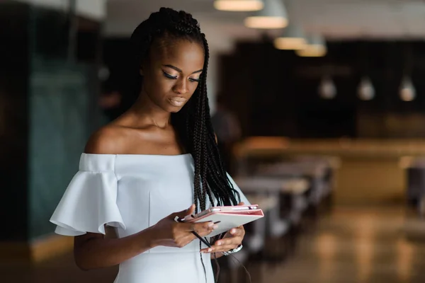 Sexy pretty Afrikaans model staat in een restaurant iets op internet zoeken met behulp van haar tablet. Meisje is het dragen van Witte avondjurk met blote schouders en horloge. — Stockfoto