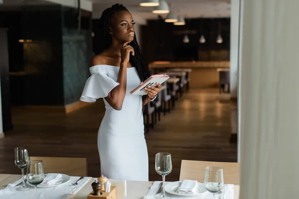 Schattig sexy vrouw met zwarte huid is stending in luxe restaurant in de buurt van goed ingerichte tabel. Meisje heeft tablet in haar hand. Model is het venster kijken en denken over iets. — Stockfoto