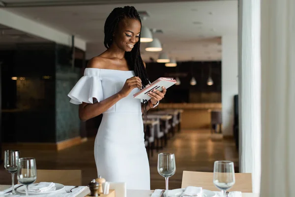 Schattig vrij sexy Amerikaanse vrouw is stending in luxe restaurant in de buurt van goed ingerichte tabel. Meisje heeft tablet in haar handen en zoeken naar de informatie op internet. Ze ziet er erg blij. — Stockfoto