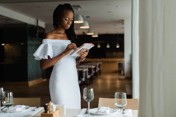 Schattig vrij sexy vrouw met zwarte huid is stending in luxe restaurant in de buurt van goed ingerichte tabel. Meisje heeft tablet in haar handen en zoeken naar de informatie op internet. — Stockfoto