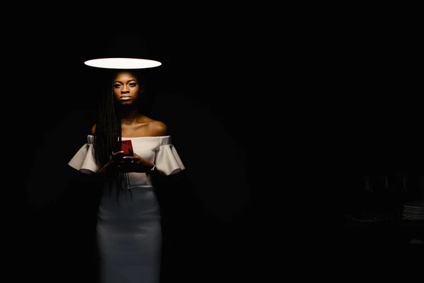 Woman in a dark room. Bright light above her head. Model is wearing white dress and has a red glass in her hands. — Stock Photo, Image