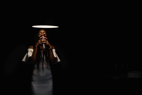 Woman in a dark room. Bright light above her head. Model is wearing white dress and drinking wine. — Stock Photo, Image