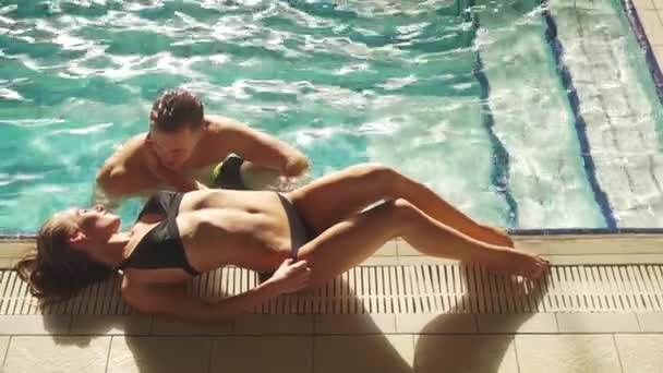 Giovane coppia nuotare in piscina in hotel di lusso, divertirsi e parlare. L'uomo sembra eccitante verso la ragazza. Donna bionda sdraiata sul bordo della piscina. Lei sta abbracciando l'uomo e sorridendo . — Video Stock