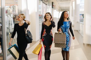 Üç charmed çekici kadın büyük alışveriş merkezinde alışveriş var. Milano ile elbise giyen kadın hortumlar ve ellerinde alışveriş torbaları. Çok mutlu ve saç ile oynarken.