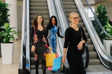 Üç arkadaş alışveriş merkezinde alışveriş var. Onlar ikinci kattan aşağı gitmek yürüyen merdiven kullanarak. Uzun elbiseler ve alışveriş torbaları ellerinde kızlar. Sarışın kız bir at kuyruğu ve gözlük vardır.