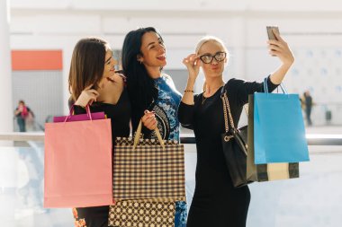 Çekici güzel kadınlar alışveriş sonra selfie alarak. Kızlar çok gülümseyen mutluyuz. Saç, yüz ve gözlük ellerine alarak fotoğraflar için poz. Birçok renkli alışveriş torbaları var..