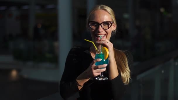 Belle fille blonde buvant un cocktail bleu avec de la glace et de la menthe, regardant la caméra et souriant . — Video