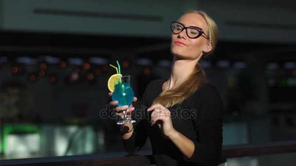Красивая девушка с хвостиком пьет синий коктейль со льдом и мятой, смотрит на камеру и солнце, улыбается. Играет со своими волосами и очками — стоковое видео