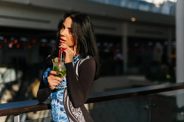 Aantrekkelijke brunette met cocktail in haar hand die verblijft in het winkelcentrum. Ze dragen elegante jurk. Als we kijken naar de camera. Mohito met ijs en munt — Stockfoto