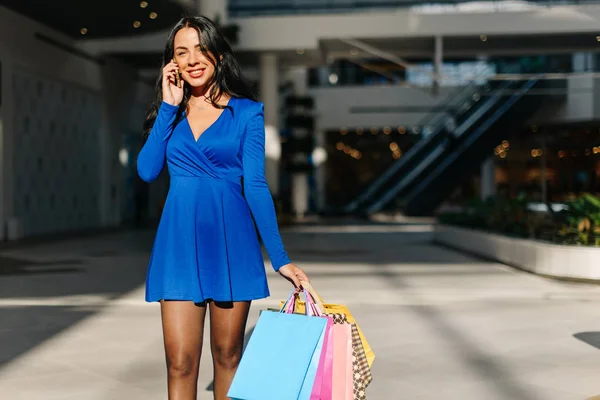 Menina agradável em vestido azul em saltos altos andando depois de fazer compras e falar por telefone. Mulher tem muitos sacos de compras na mão . — Fotografia de Stock