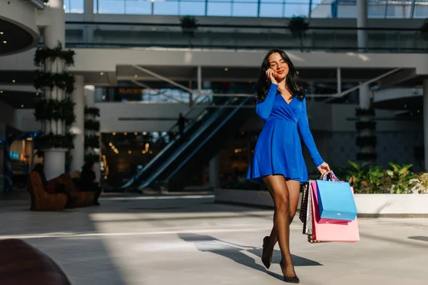 Menina bonita em vestido azul com mangas compridas em saltos altos depois de fazer compras e falar por telefone. Mulher tem muitos sacos de compras coloridos em sua mão . — Fotografia de Stock