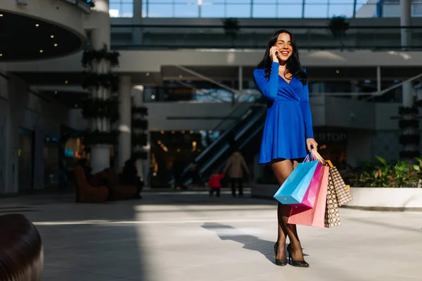 Mulher atraente em vestido azul com mangas compridas em saltos altos pretos depois de fazer compras, falando por telefone. Mulher tem muitos sacos de compras coloridos em sua mão. Centro comercial cheio de luz solar — Fotografia de Stock