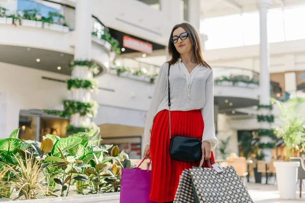 Szép barna lány után shopping mall. Vonzó nő visel piros midi szoknya, fehér ing, poharak. Bevásárló szatyrok van a kezében. Tartózkodó mall, és mosolyogva — Stock Fotó