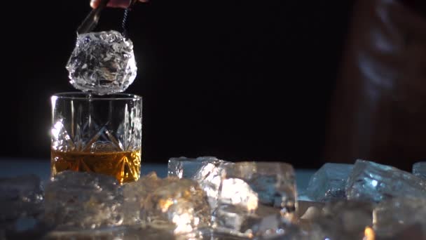 Bartender taking a ball of ice into glass with wiskey. Drink is spleshing on the sides. Dark and shadows — Stock Video