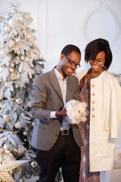 Het mooie Afro-Amerikaanse echtpaar is blij dat het kleine pluizige witte konijn in de buurt van de kerstboom op de Happy New Year Eve — Stockfoto