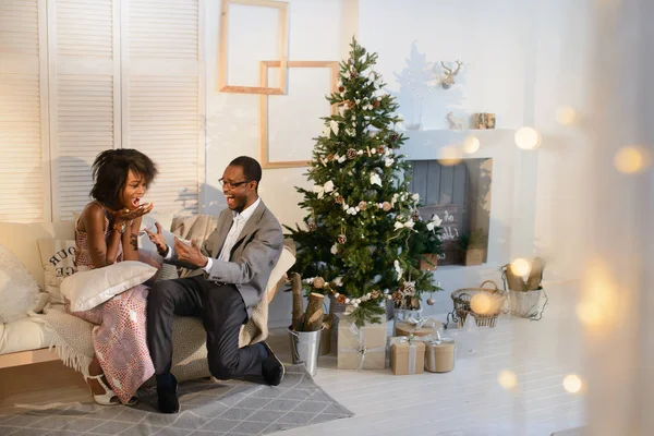 O homem afro-americano atraente bonito está fazendo uma grande surpresa para sua namorada na véspera de Natal, ela está muito surpreendida e não consegue lidar com emoções. — Fotografia de Stock