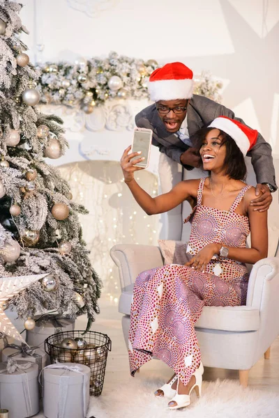 O belo casal afro-americano em bonés de Natal fazendo selfies e celebrando a véspera de Natal perto da árvore de Natal — Fotografia de Stock