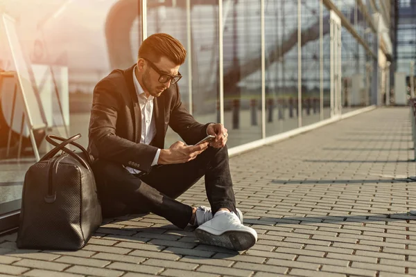 Pohledný vousatý podnikatel ve stanici letiště psaní textové zprávy na jeho mobilní telefon a sedí na zemi vedle vchodu — Stock fotografie