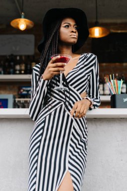 Oldukça çekici Amerikalı kadın elinde kokteyl kırmızı çubuk. Çizgili uzun elbiseli güzel kadın. Duvar üzerine alkol şişe dolu. Birçok payet bar kokteyller için. Buz kokteyller.