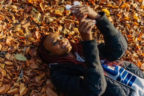 Krásná africká americká dívka ležící v žluté listí v podzimním parku. Ženské tvorby selfi a usmívá se — Stock fotografie
