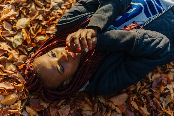Linda menina afro-americana deitada em folhas amarelas no parque de outono. Feminino fazendo selfi e sorrindo — Fotografia de Stock