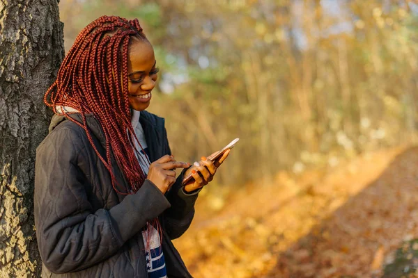 Hezká žena stojící poblíž stromu v podzimním parku při západu slunce. Dívka v Internetu pomocí telefonu a usmívá se. Krásný autamn park — Stock fotografie