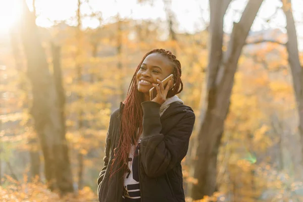 Linda chica afroamericana tiene una conversación en el parque de otoño. Las mujeres descansan al aire libre y el humor bueno. La hembra sonríe — Foto de Stock