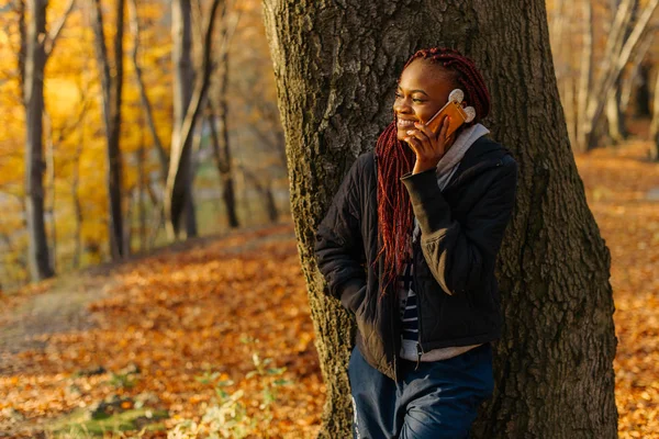 Mujer hablando por teléfono en el parque de otoño al atardecer. Niña de pie cerca del árbol y mirando las hojas amarillas. Hermoso parque autamn — Foto de Stock