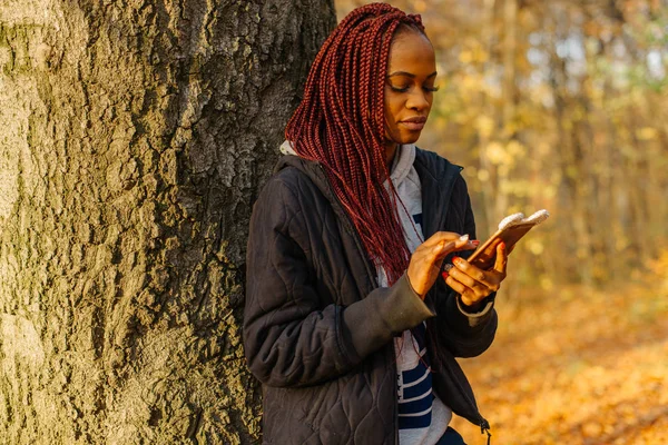 Žena stojící poblíž stromu v podzimním parku při západu slunce. Dívka hledání něco v telefonu. Krásný autamn park — Stock fotografie