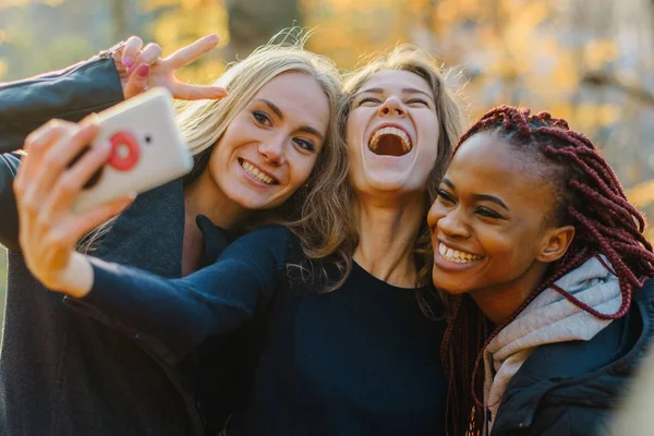 Três mulher bonita fazendo selfie no parque de outono. Meninas bonitos com pele de cor diferente. Feminino fazendo caras engraçadas e sorrindo para a câmera — Fotografia de Stock