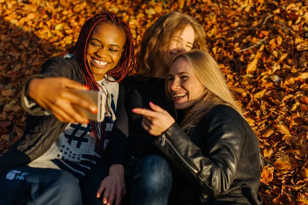 Drei Frauen machen im Herbstpark ein lustiges Selfie. niedliche Mädchen mit unterschiedlich gefärbter Haut. Weibchen liegen in gelben Blättern und machen lächelnde Gesichter in die Kamera. sonniger Herbsttag — Stockfoto