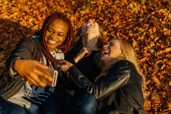 Amigos femininos muito felizes fazendo selfie engraçado no parque de outono. Meninas bonitos com pele de cor diferente. Mulher deitada em folhas amarelas e fazendo rostos sorridentes para a câmera. Dia de outono ensolarado — Fotografia de Stock