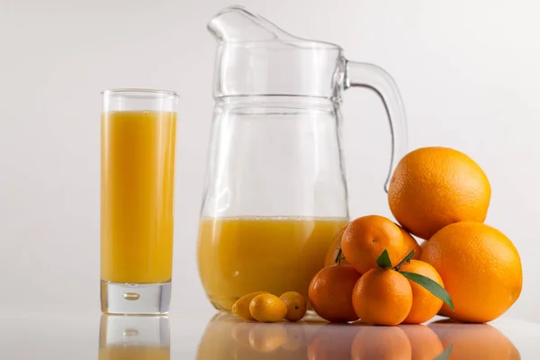 Composition du verre plein de jus d'orange, quelques oranges et mandarines et un pot sur une surface blanche — Photo