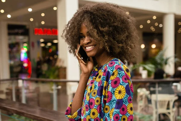 Афро-американка разговаривает по телефону и улыбается, стоя в торговом центре — стоковое фото