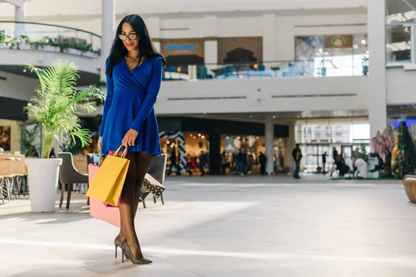 Atraente tímida mulher feliz em vestido azul curto com mangas compridas em saltos altos pretos e óculos andando depois de fazer compras. Feminino tem muitos sacos de compras coloridos em suas mãos . — Fotografia de Stock