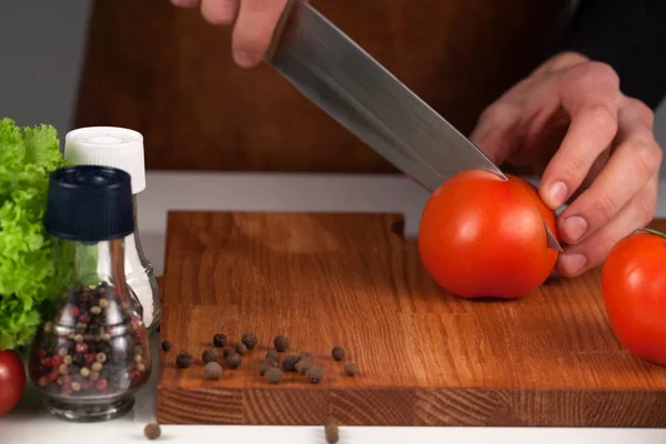 Γκρο πλαν του σεφ χέρια κοπής κόκκινη ντομάτα σε ένα ξύλινο κοπή του σκάφους. Τοποθετούνται με το μαρούλι, το πιπέρι και το αλάτι — Φωτογραφία Αρχείου