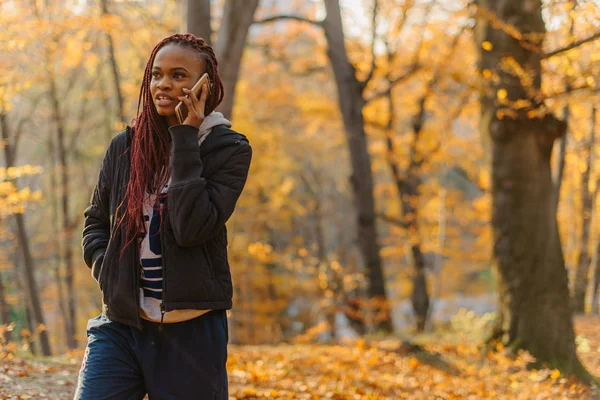 Linda chica afroamericana caminando en autamn park y hablando por teléfono. La mujer descansa al aire libre . — Foto de Stock