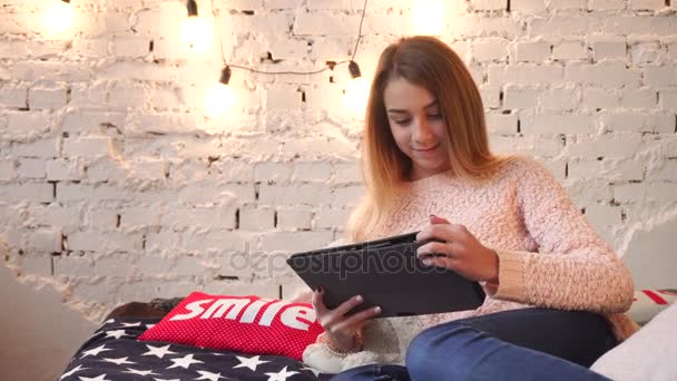Krásná mladá dívka chatování na internetu na pohovce s měkkými polštáři a věnec na zeď. — Stock video