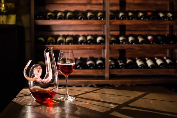 Composition af carafe de vin et verre à vin placés sur une table en bois. Étagères avec bouteilles de vin sur le fond . — Photo