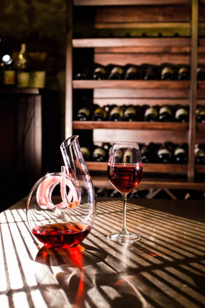 График вина и бокал вина, наполненный красным вином, помещенным на деревянный стол. Полки с бутылками вина на заднем плане . — стоковое фото