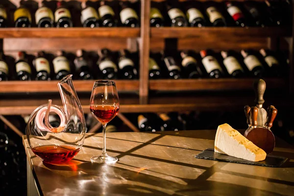 Composition of wine carafe, wine glass and cheese placed on a wooden surface. Shelves with wine bottles on the background. — Stock Photo, Image