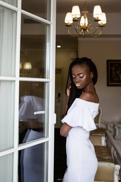 Sottile ragazza afro-americana in posa in bellissimo vestito bianco in una grande stanza vicino alla finestra . — Foto Stock